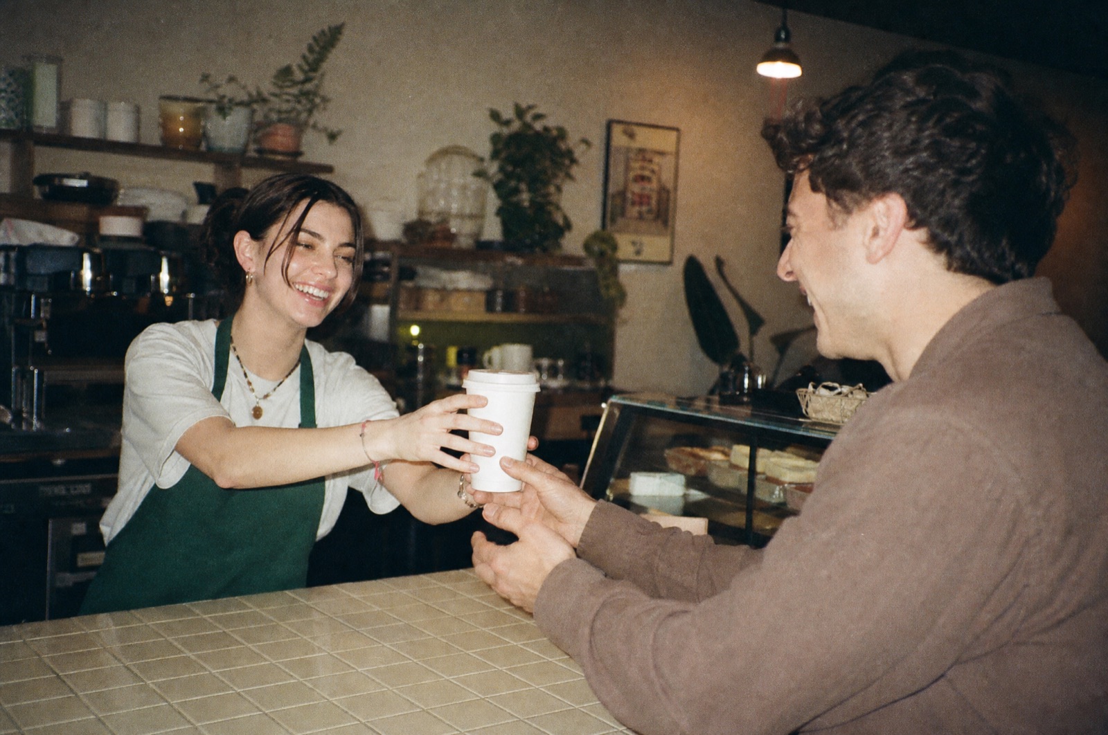 A smiling barista in a green apron handing a coffee cup across the counter directly into a regular's hands