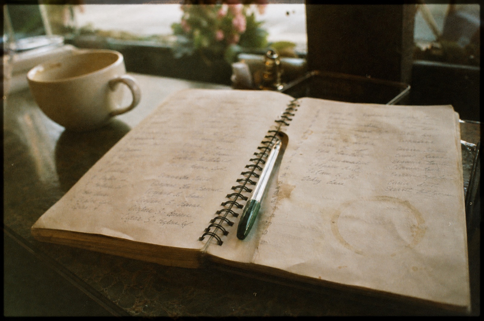 A worn spiral notebook of handwritten regulars' names lying open on a café counter beside a cup of coffee