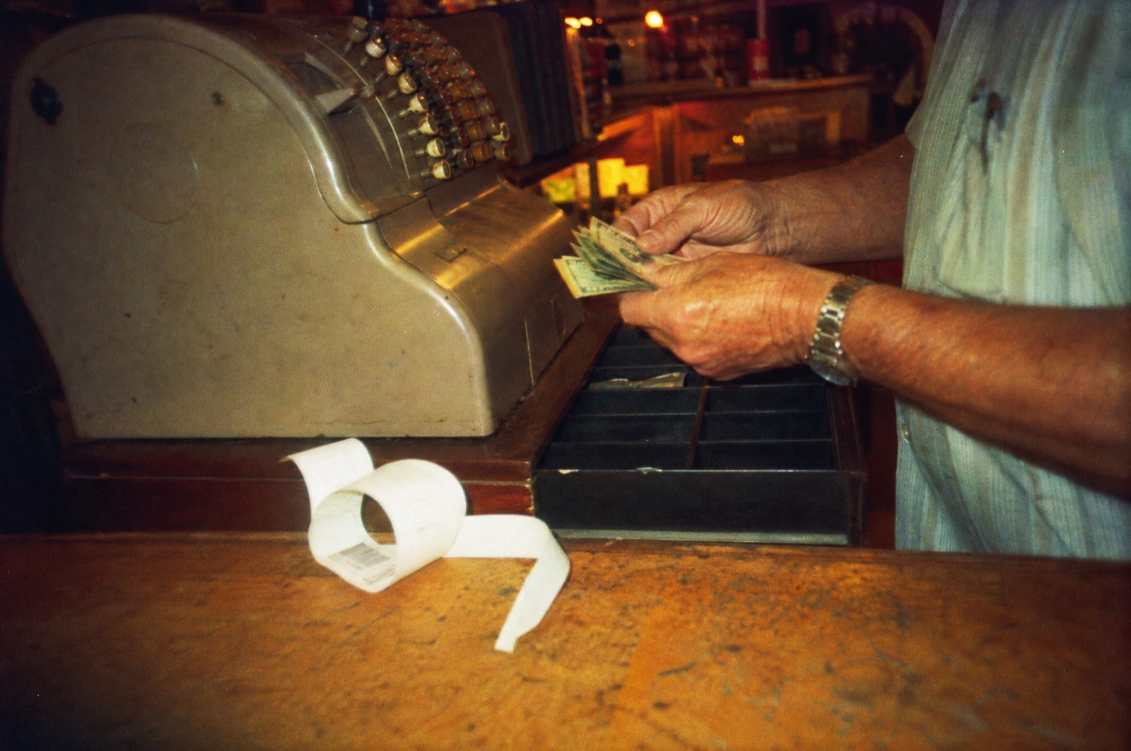 Weathered hands counting a small stack of bills over the open drawer of an old cash register