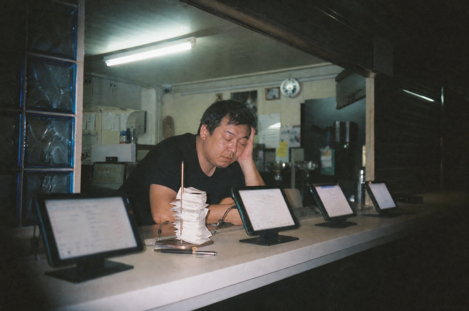 An exhausted restaurant owner late at night, head in hand behind a counter lined with four glowing order tablets and a spike of receipts