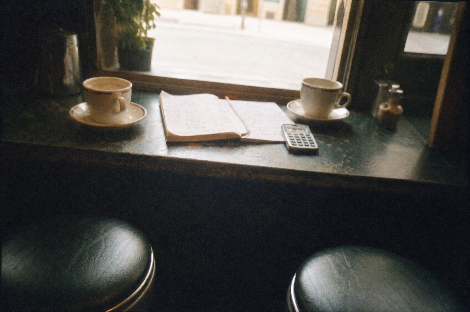 Two cups of coffee on a worn café window counter beside an open notepad of handwritten arithmetic and a pocket calculator