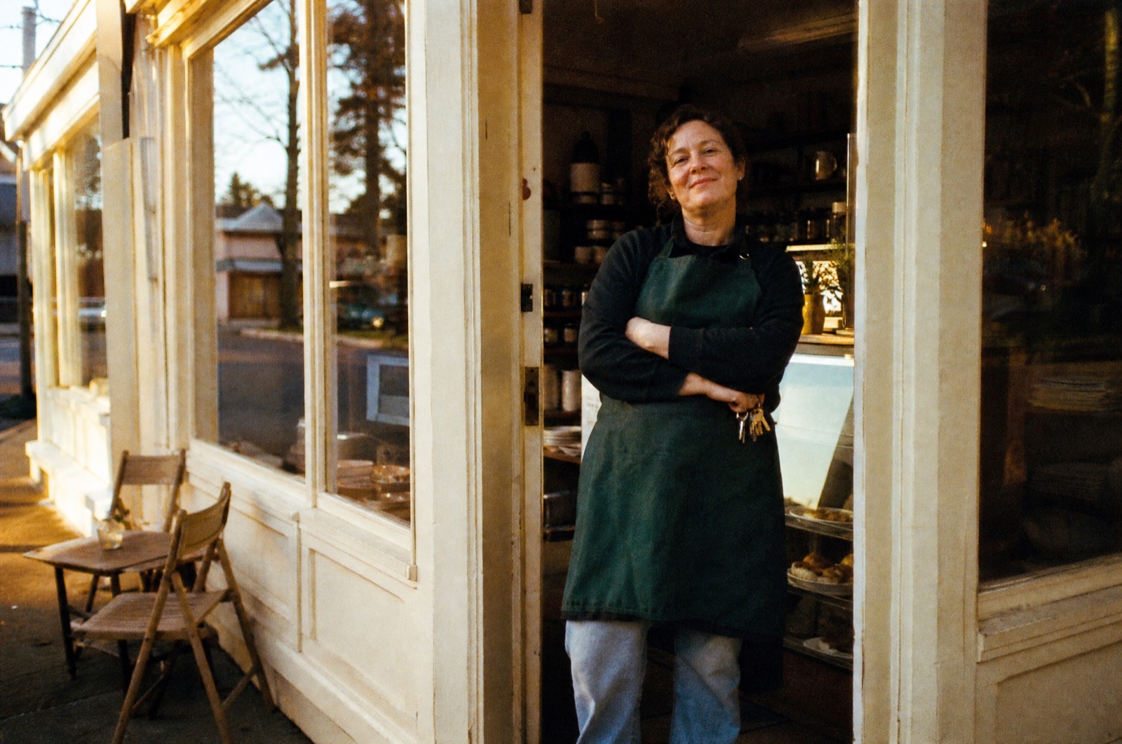 A café owner in a deep green apron standing proudly in her shop doorway at golden hour, keys hooked on one finger